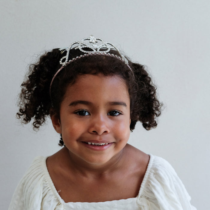 A young girl with curly brown hair wearing a silver tiara and a white dress, photographed against a plain light gray background.
