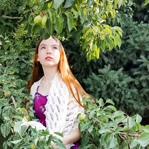 Woman with long red hair wearing a white lace knit shawl over a purple dress standing among green foliage and pear trees in a garden