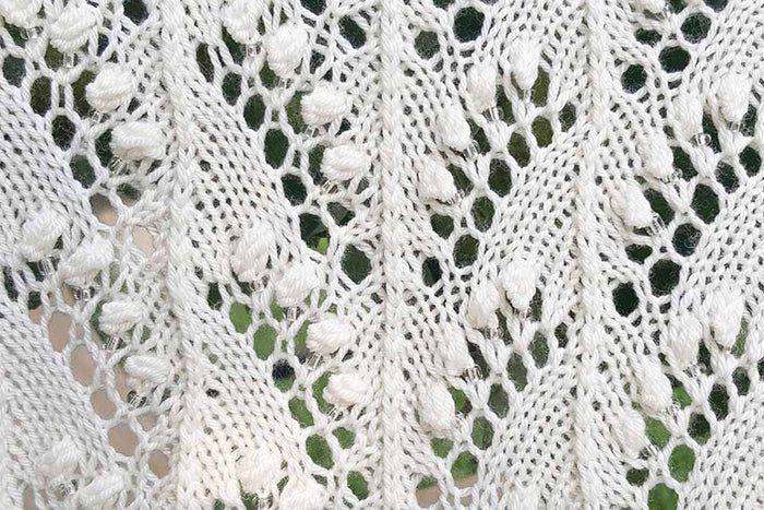 Detail of white lace shawl showing yarn-over eyelets and leaf motifs in the border pattern against a white background