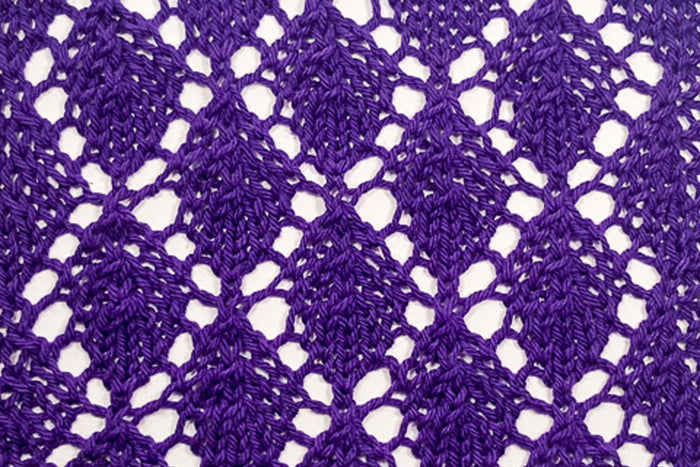 Close-up of a lace knitting stitch pattern sample in deep purple yarn showing an open diamond lattice pattern from the Conquering Lace class