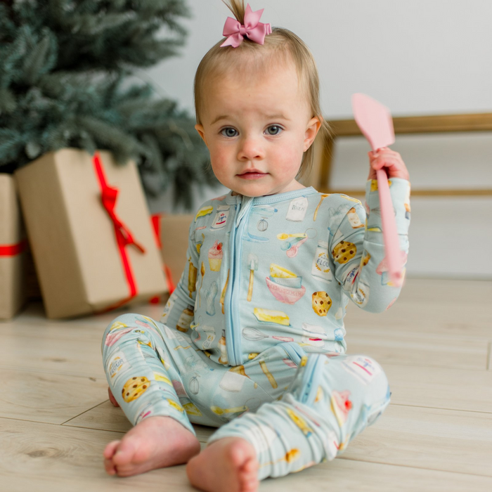 baby girl sitting on wood floor holding spatula wearing blue zip romper with multi color baking themed print all over