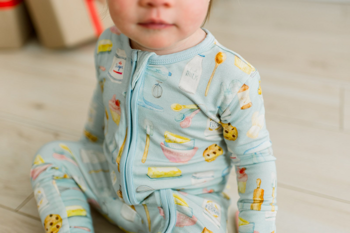 baby sitting on wood floor wearing blue zip romper with multi color baking themed print all over