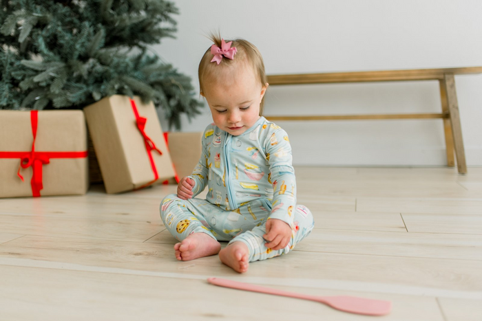 baby girl sitting on wood floor with spatula wearing blue zip romper with multi color baking themed print all over