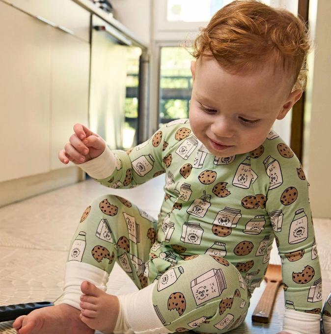 A baby with red hair sitting on a wooden floor wearing a light green one-piece pajama with a cookie and milk carton pattern.