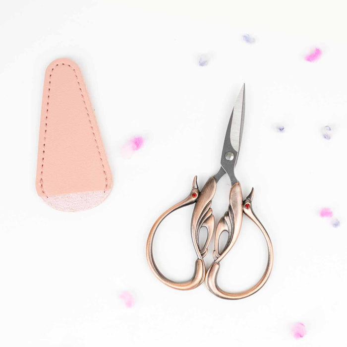 Copper swan embroidery scissors with ornate swan-shaped handles and red crystal eyes next to a pink leather blade cover with scattered yarn bits