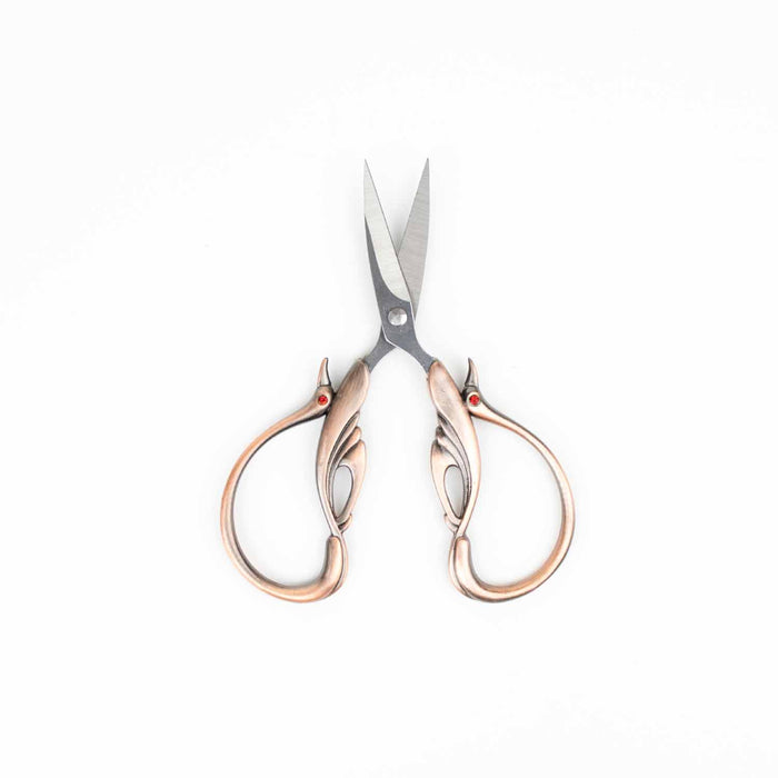 Copper swan embroidery scissors standing upright with blades pointing up showing the symmetrical swan wing handle design with red crystal eyes