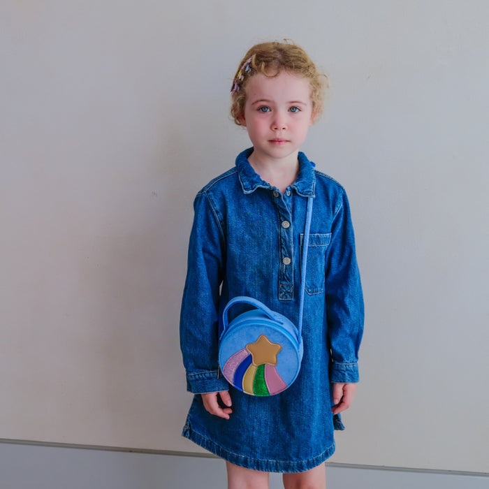 A young child wearing a blue denim shirt dress and holding a round blue crossbody bag with a colorful star design, standing against a plain light gray wall.