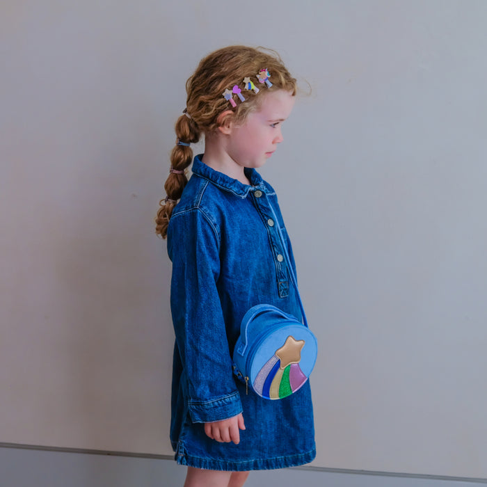 A young girl with a long braid is shown in profile wearing a blue denim shirt dress and carrying a round blue crossbody bag with a rainbow and star design. She is standing against a plain light gray wall.