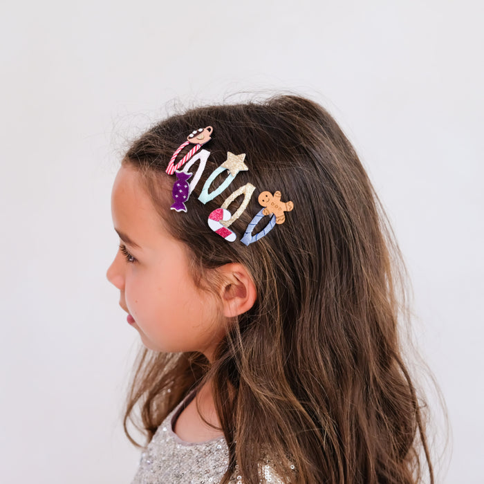 A side-profile view of a young girl with long brown hair wearing a colorful set of three hair clips. The clips feature pastel-colored designs including a purple mermaid tail, a gold star, and a gingerbread cookie, set against a plain light gray background.