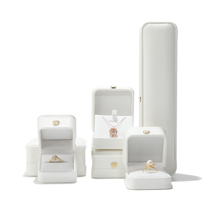 A sophisticated set of white leather jewelry boxes with gold crown emblems, showcasing a diamond ring, a pearl ring, and a rose gold necklace on a clean, high-contrast background.