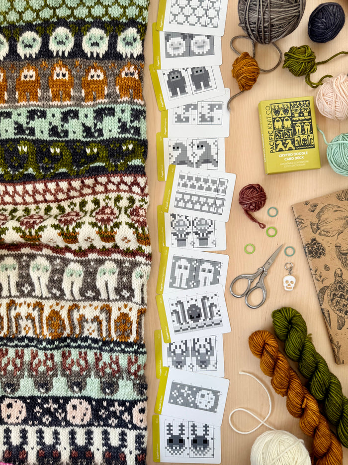 Cryptid Doodle chart cards beside a monster-motif colorwork knit and mini skeins