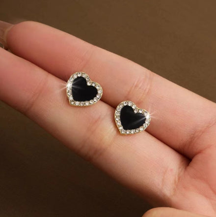 A high-resolution product shot featuring two pairs of heart-shaped stud earrings; one pair is silver with an open-heart design encrusted in sparkling crystals, and the other is gold-toned with a solid black onyx-style center surrounded by a crystal halo, both displayed against a pure white background.