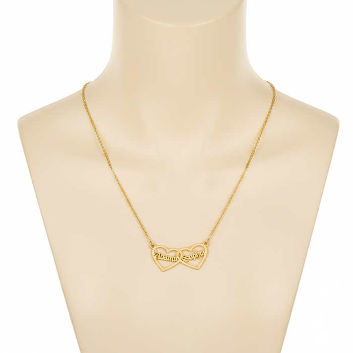 A gold-toned necklace featuring two interlocking open-heart pendants, displayed on a beige mannequin bust. Each heart is personalized with a name in a Gothic-style font: "Alonma" on the left and "Sophia" on the right. The pendant is attached to a fine gold cable chain.
