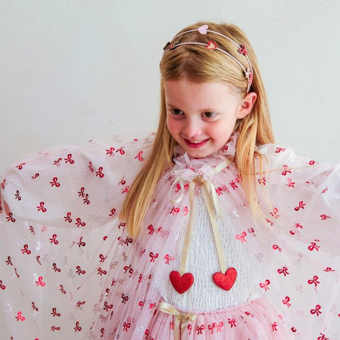 A young girl with blonde hair wearing a pink and red bow-patterned tutu dress with a white knit bodice, a cream ribbon tie, and a matching sheer pink headband with heart details.