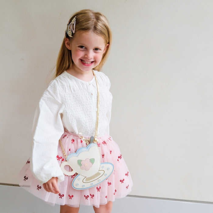 A young girl with blonde hair wearing a white long-sleeve top and a pink tulle skirt with red bow prints, smiling while holding a decorative teacup-shaped crossbody bag.