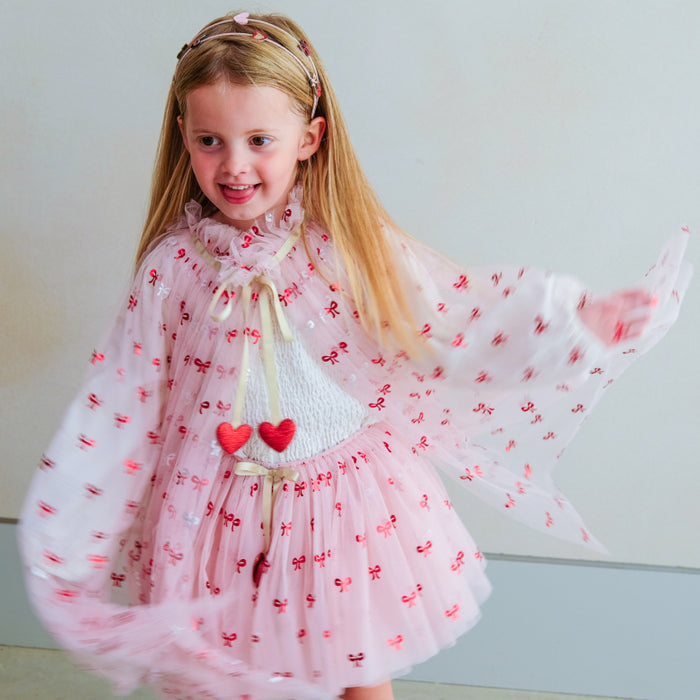 A young girl with long blonde hair wearing a pink tulle dress with red bow patterns, a white knit bodice, and a sheer pink overlay, standing against a plain light gray wall.