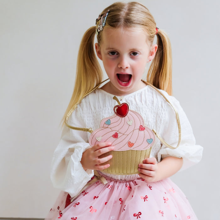 A young girl with blonde pigtails wearing a white blouse and a pink tutu skirt, holding a pink and gold cupcake-shaped purse with a red heart on a plain light gray background.