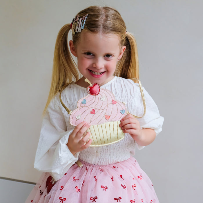 A young girl with blonde pigtails smiles while holding a cupcake-shaped purse. She is wearing a white long-sleeve blouse and a pink tulle skirt with small bow prints.