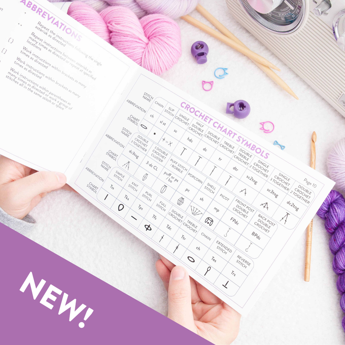 Hands holding open Dawn's Handy Reference Guide showing the crochet chart symbols page with tables of abbreviations and stitch diagrams
