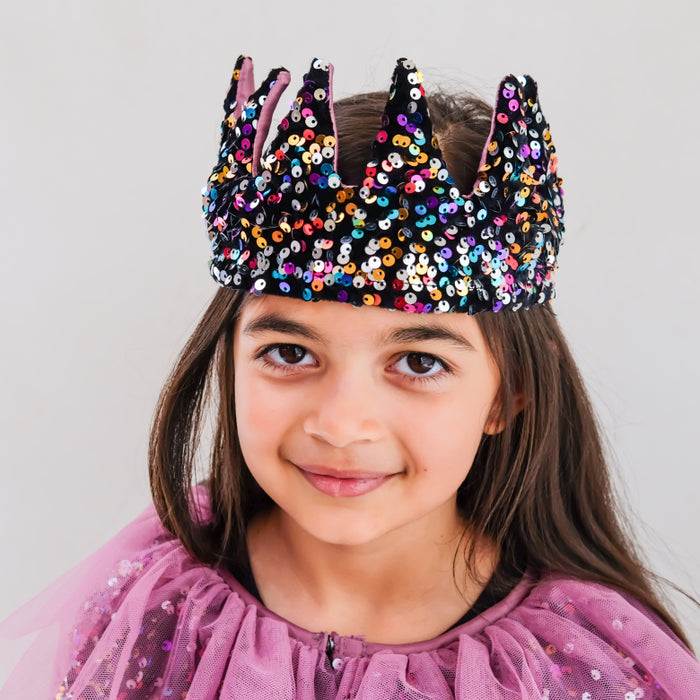 A young girl with brown hair wearing a black sequined crown and a purple tulle dress, photographed against a light gray studio background.