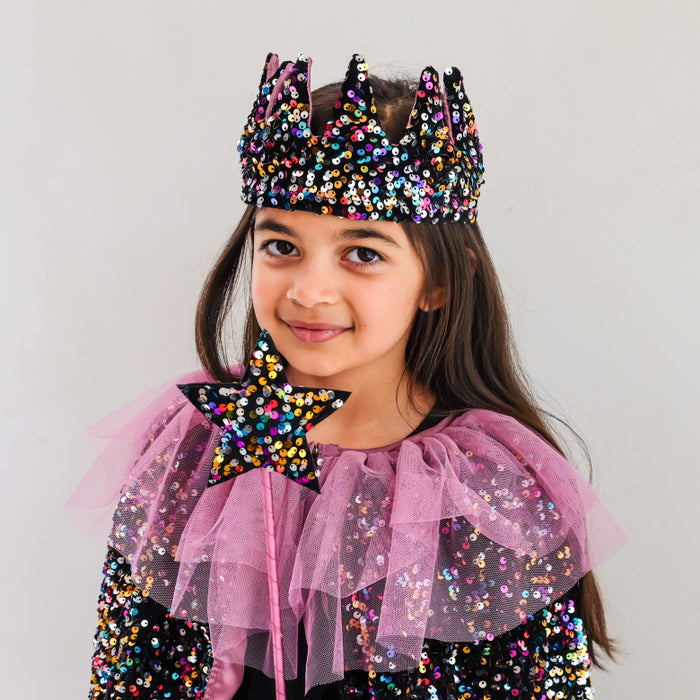 A young girl wearing a black sequined crown with multicolored glitter, a pink tulle ruffled collar, and a matching star-shaped accessory, photographed against a plain light background.