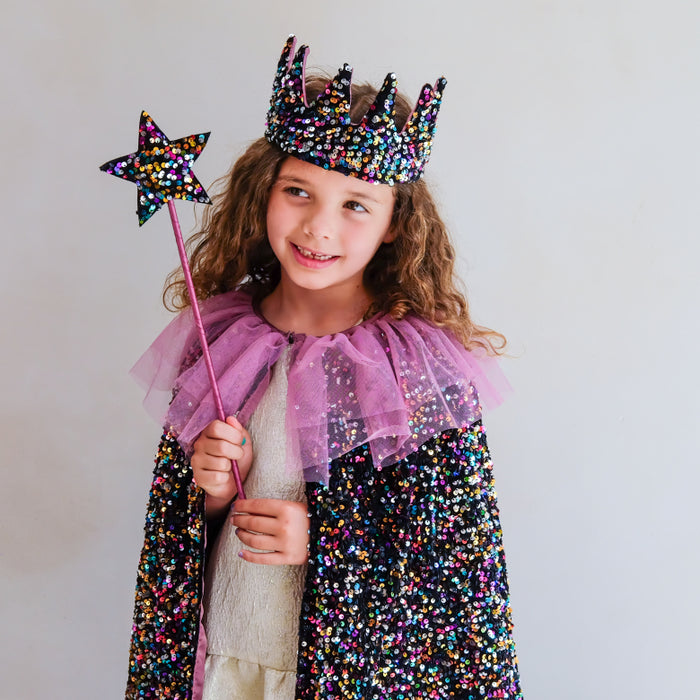 A young girl wearing a black sequined royal cape with a purple tulle collar, a matching black sequined crown, and holding a star wand, set against a plain light gray background.