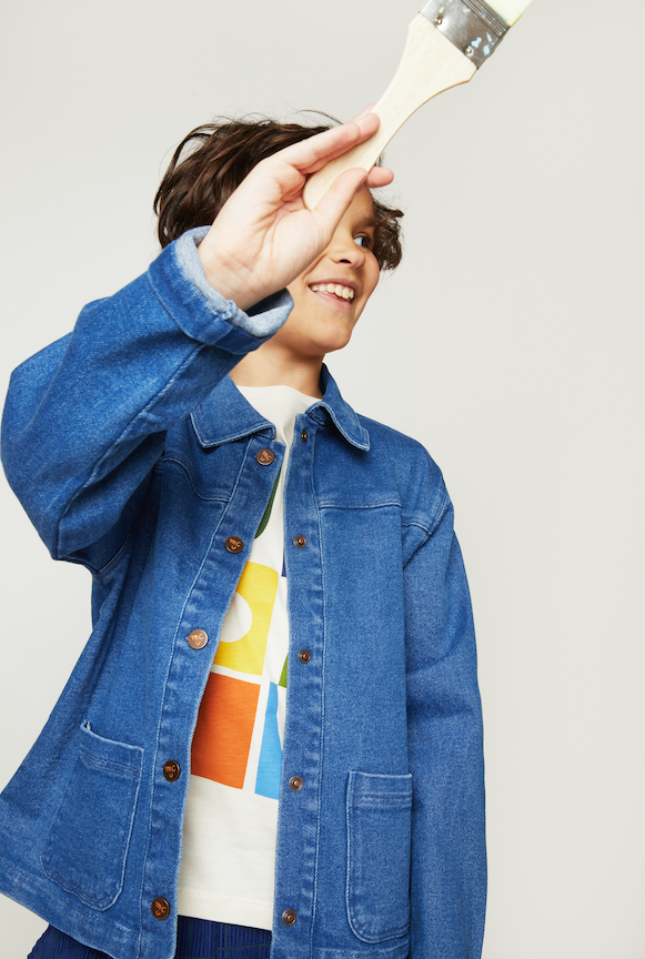 A child wearing a blue denim jacket over a white graphic t-shirt, holding a paintbrush to their eye against a light gray studio background.