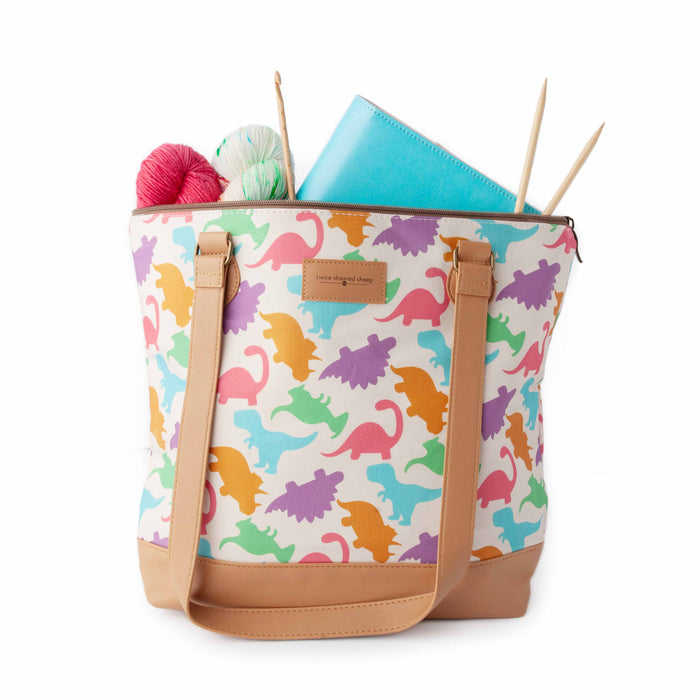 Dino Mega Trinity Bag open with yarn skeins in pink and mint green, a blue project journal planner, and bamboo knitting needles peeking out the top