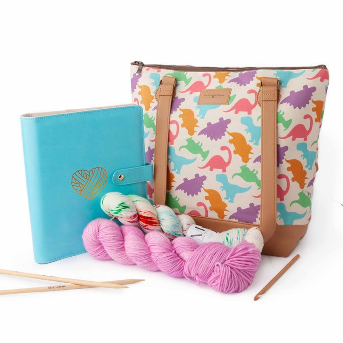 Dino Mega Trinity Bag styled with a blue project journal planner with yarn heart emblem, pink and speckled yarn skeins, bamboo knitting needles, and a crochet hook arranged in front
