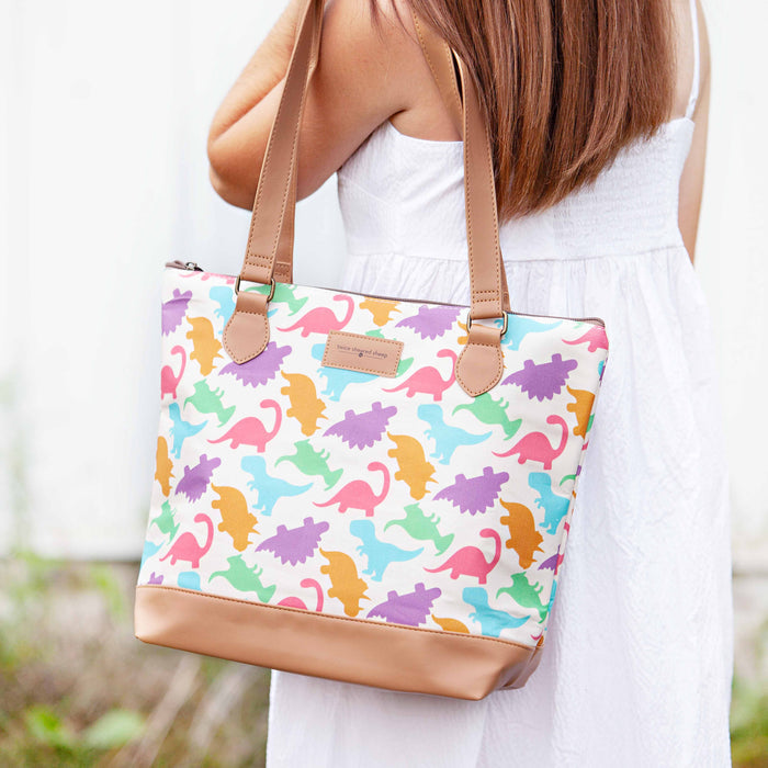 Person carrying the Dino Mega Trinity Bag on one shoulder showing the colorful dinosaur silhouette print canvas with tan vegan leather bottom and strap against a white dress