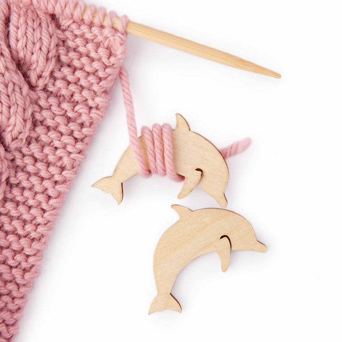 Wooden dolphin yarn bobbins wrapped with pink yarn next to a knitting project on a wooden needle