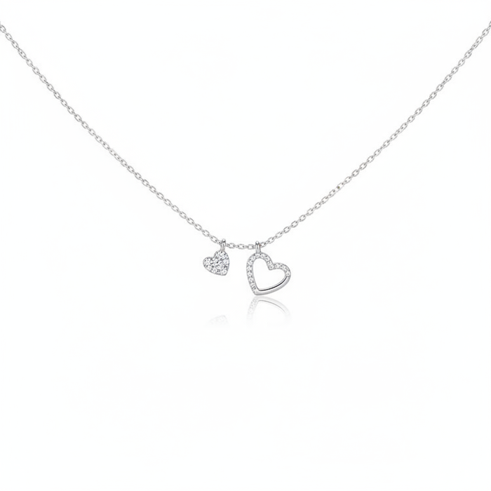 A delicate silver-toned necklace featuring two heart charms suspended from a fine cable chain. One charm is a small solid heart fully encrusted with pavé crystals, and the other is a slightly larger open-heart frame also lined with shimmering crystals. The jewelry is shown against a minimalist white background with a subtle reflection.