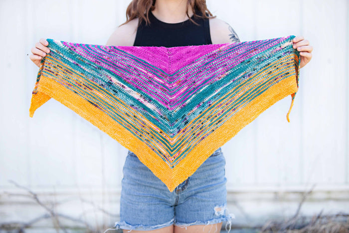 Model holding the Down By The Sea shawl fully extended, displaying the triangular shape and chevron stripe pattern in magenta, teal, and golden orange