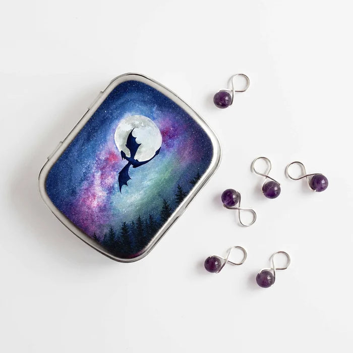 Small hinge-style Dragon Moon storage tin next to five purple amethyst infinity stitch markers, showing watercolor dragon and moon galaxy artwork on lid