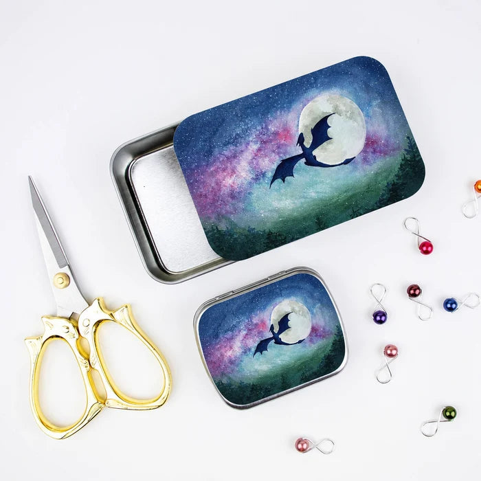 Large and small Dragon Moon storage tins styled with gold cat scissors and colorful infinity stitch markers, showing watercolor dragon moon galaxy artwork