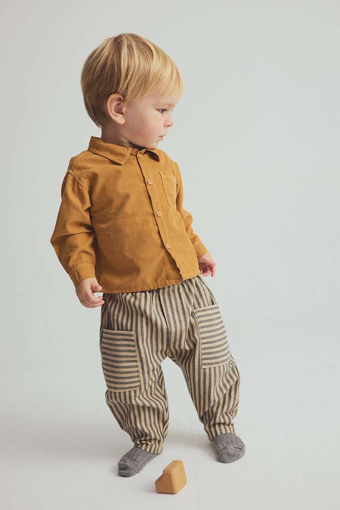 A toddler standing in a studio wearing a mustard yellow corduroy button-up shirt, striped beige and brown pants, and gray socks, with a small wooden block on the floor.