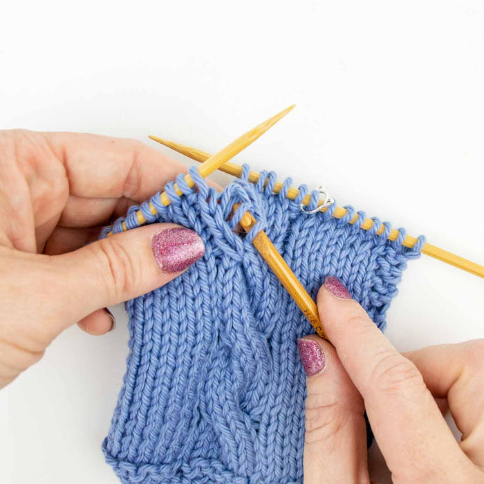 Close-up of hands using a Duo wooden repair hook to pick up a dropped stitch in blue cable knit fabric on bamboo needles