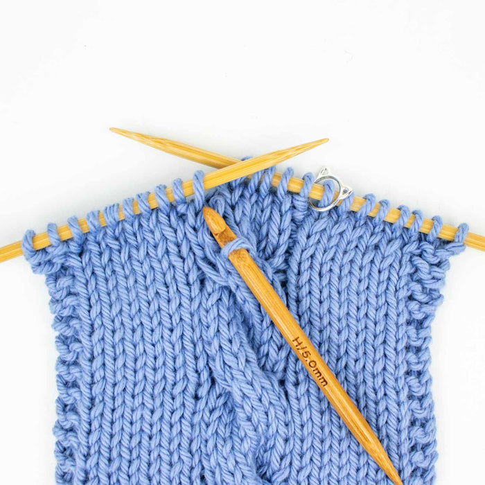 Close-up of a Duo wooden repair hook inserted into blue knit fabric on bamboo needles showing the hook catching a dropped stitch