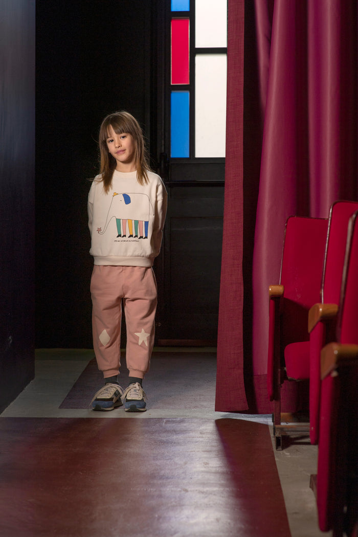 A young girl stands in a dimly lit indoor space wearing a cream sweatshirt with a colorful graphic and pink pants. Red curtains and red theater-style seats are visible in the background.