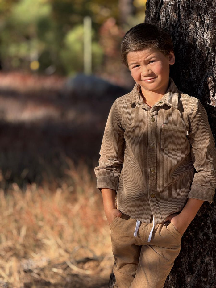 Young boy in a brown shirt and pants standing next to a tree in a natural setting