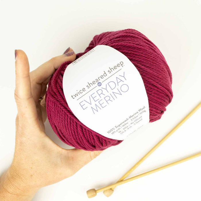 Hand holding a ball of Everyday Merino worsted weight yarn in Sugar Beet (deep pink) with Twice Sheared Sheep label and wooden knitting needles