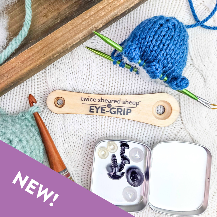 Twice Sheared Sheep Eye Grip wooden safety eye tool on white knit background with blue amigurumi in progress on green needles, wooden crochet hook, and tin of safety eyes