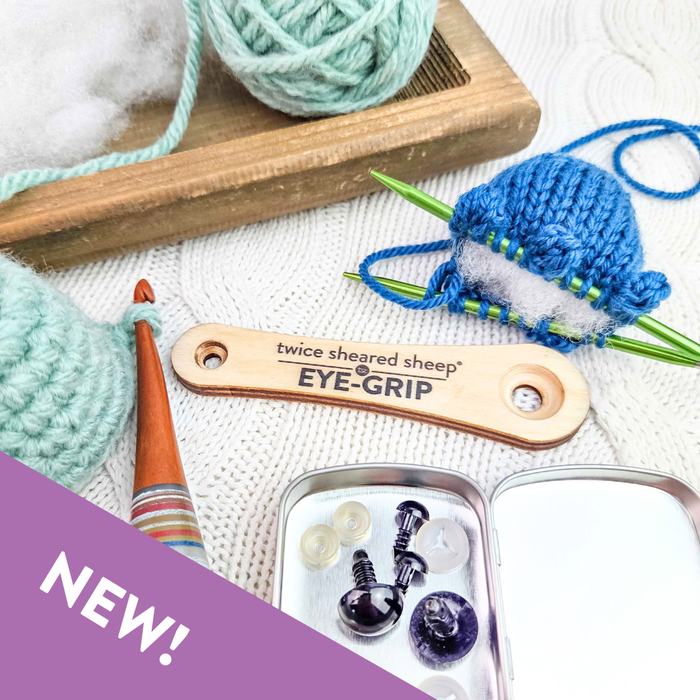 Eye Grip wooden safety eye tool styled with blue amigurumi on green needles, wooden crochet hook with mint crochet piece, safety eyes tin, and yarn in wooden tray