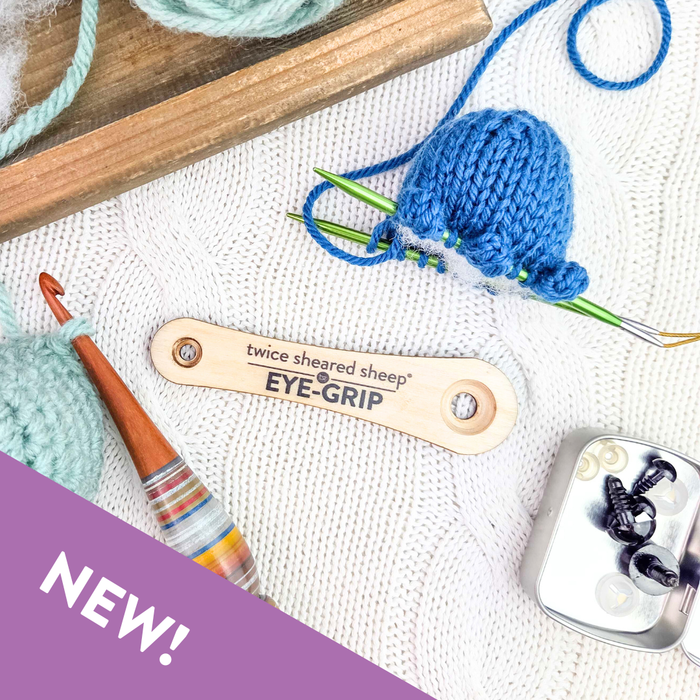 Eye Grip wooden safety eye tool on white knit background with blue amigurumi in progress, wooden crochet hook, safety eyes tin, and mint yarn at a wider angle