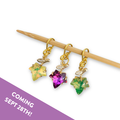 Three gold fall leaf stitch markers set with yellow, magenta and green crystals hanging from a bamboo knitting needle, with a Coming Sept 28th banner