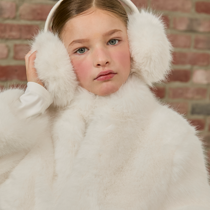 Child wearing white fur coat and ear muffs against a brick wall.