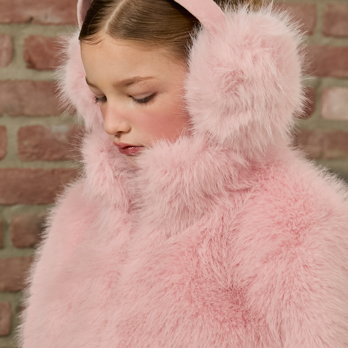 Person wearing a pink fluffy coat with earmuffs against a brick wall.