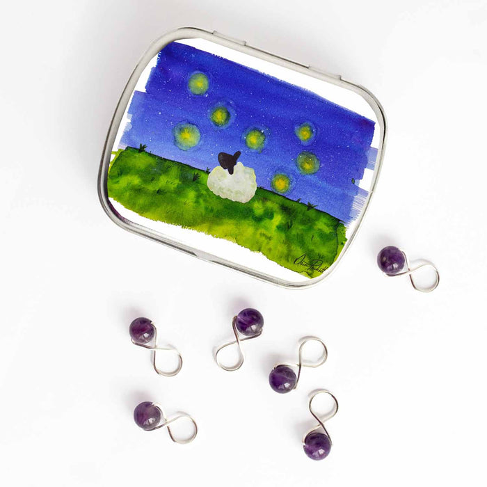 Small hinge-style Firefly Dreams storage tin open beside purple amethyst bead infinity-loop snag-free stitch markers on white background