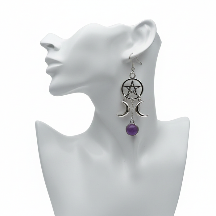 A silver-toned gothic-style dangle earring featuring a pentagram within a circle at the top. Two crescent moons hang from the sides, flanking a central chain that drops down to a round, purple amethyst-style cabochon. The earring is displayed on a white mannequin head against a plain white background.