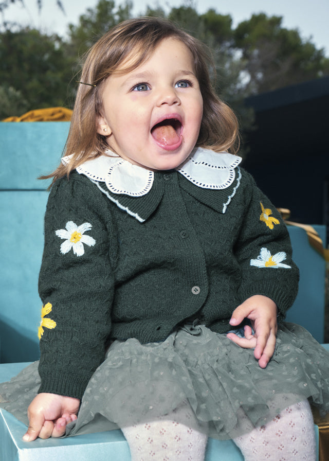 Child wearing a green cardigan with floral designs, sitting outdoors.